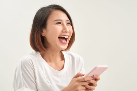 Cheerful asian woman standing isolated over white background, holding mobile phone, celebrating