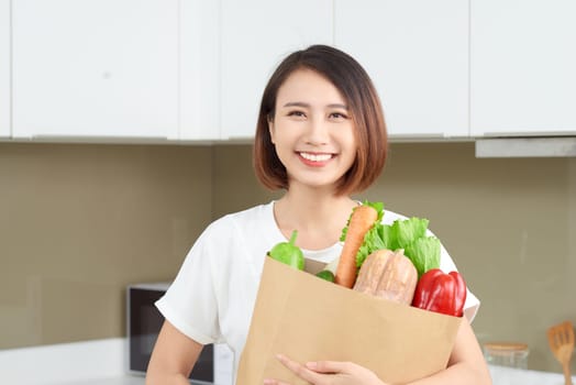 Happy, cheerful Asian woman holding paper bag full of fresh products, vegetables, greens, baguette and bananas.