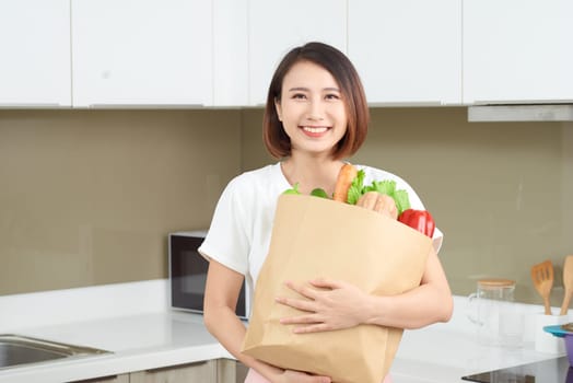 Young Asian woman holding grocery shopping bag with vegetables in the kitchen.