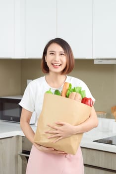 Happy, cheerful Asian woman holding paper bag full of fresh products, vegetables, greens, baguette and bananas.
