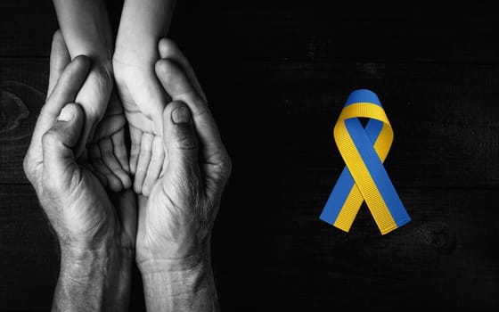 male hands hold childrens on dark wooden background with yellow and blue ribbon. black and white color. concept needs help and support, truth will win