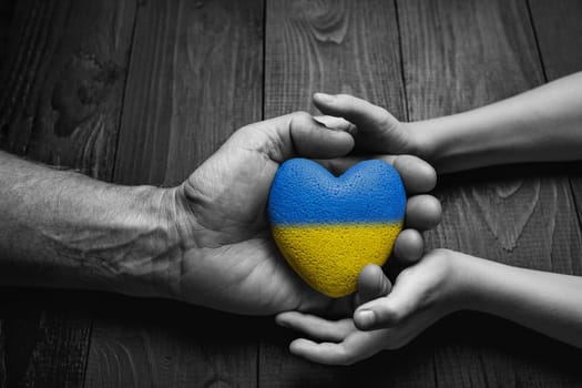 male hand gives yellow and blue heart to childs hand on dark wooden background. black and white color. concept needs help and support, truth will win