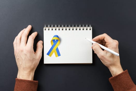 I am writing letter, I have pen in my hands and yellow blue ribbon on notebook on dark backgroun. concept needs help and support, truth will win