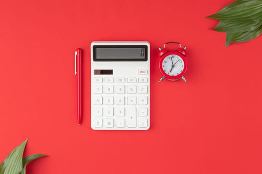 White calculator with a pen, alarm clock and green leaves on a red background. Top view.