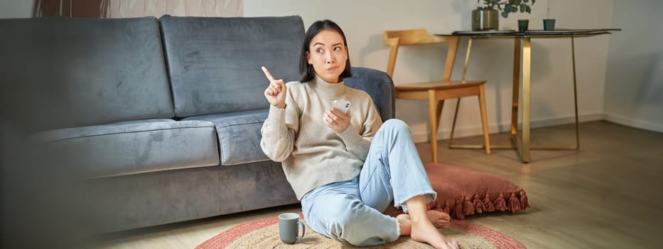 Portrait of woman sitting on floor with smartphone, looking thoughtful and pointing finger at banner, promo advertisement on top right corner.