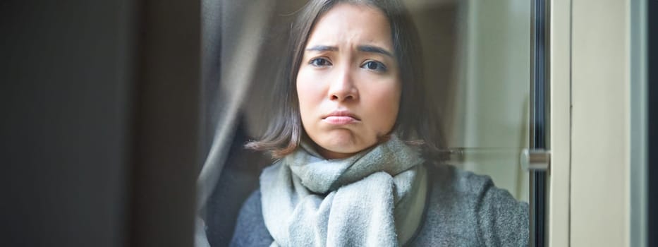 Portrait of sad ill woman staying at home, looking out of the window with jealous and upset face, cant leave home as being sick with covid-19.