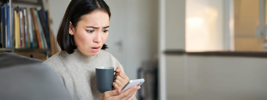 Close up of asian girl with cup in hand, looking shocked at mobile phone screen, reading concerning news on smartphone app.