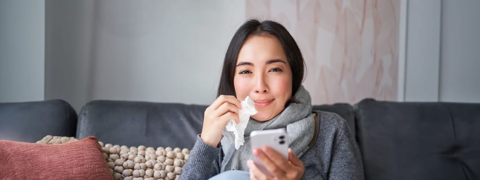 Portrait of sick woman contacting her GP via mobile phone while catching cold, sitting with flu at home, using smartphone.