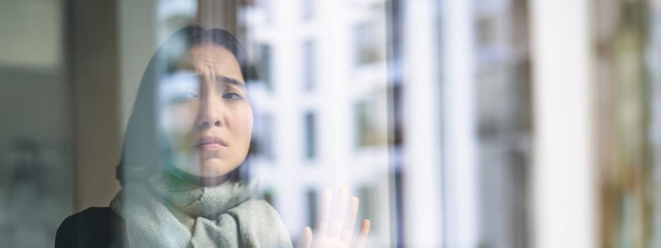 Asian woman feels unwell, stays at home, looks outside window with jealous, distressed face, wants go outside but caught cold.