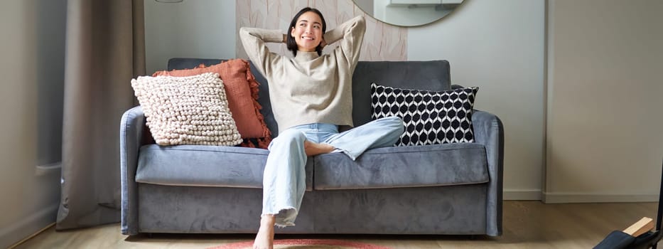 Portrait of happy asian woman feeling lazy, stretching on sofa and smiling pleased, relaxing at home, resting from work.