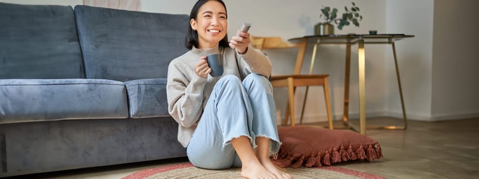 Image of young korean woman drinks coffee, holds remote from tv, watching television at home, resting in her house.