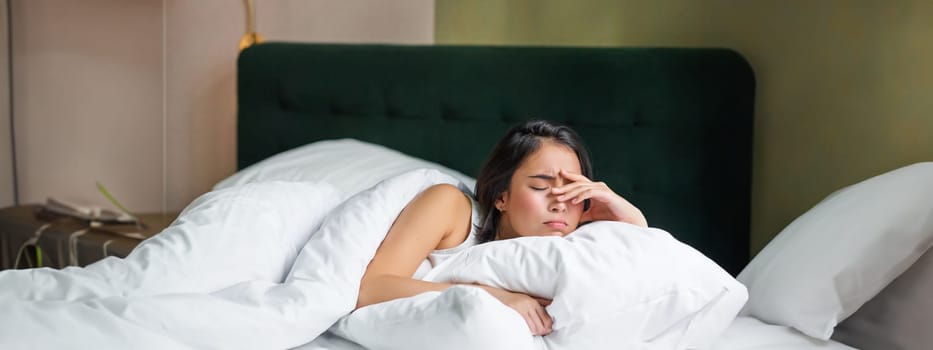 Sad and distressed young korean woman lying in her bed with pillow and warm duvet, feels uneasy, frowning and grimacing.