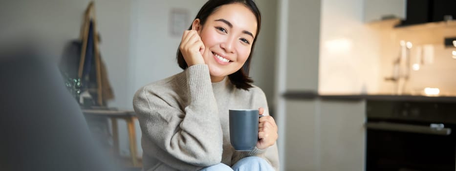 Smiling asian woman sitting at home with cup of coffee, relaxing and feeling warmth, looking outside window, resting on sofa in living room.