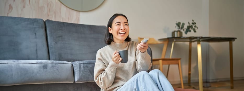 Portrait of beautiful asian girl sitting at her home and watching tv, holding remote, smiling and laughing, feeling comfort and warmth at her apartment.