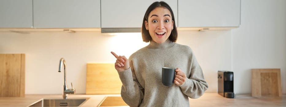 Portrait of smiling asian girl standing in kitchen, drinking coffee from cup and pointing at banner, showing letting agencies advertisement.