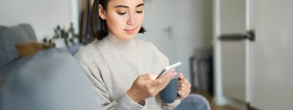 Portrait of smiling asian girl checking her news feed on smartphone and drinking coffee, sitting on sofa at home, browsing on mobile phone, reading.