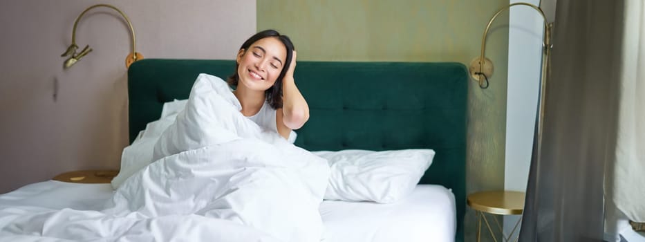 Happy korean woman wakes up in her hotel room, lying in bed under cozy warm blankets, white sheets, enjoying weekend morning.