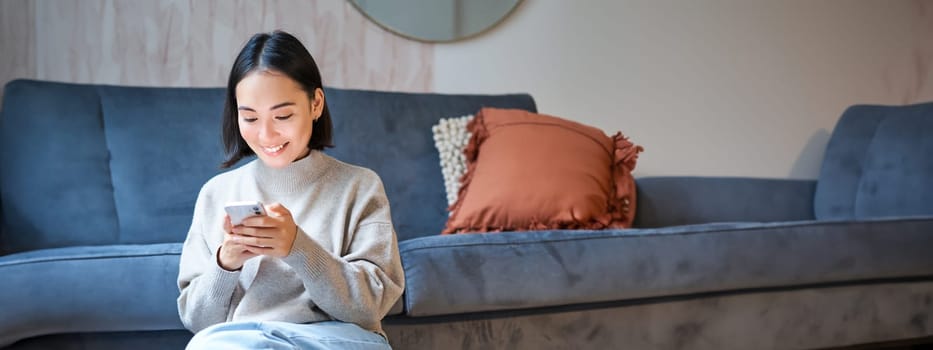 People and technology. Smiling asian woman sitting at home, using her mobile phone, typing message, browing internet or shopping online from smartphone app.