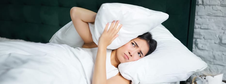 Insomnia. Asian girl shuts her ears with pillow as partner snoring. Woman lying in bed, bothered by noise at night, cannot sleep.