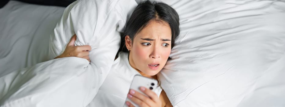 Close up portrait of asian girl lying in bed, looking at smartphone concerned, waking up late and staring at her alarm clock on mobile phone.