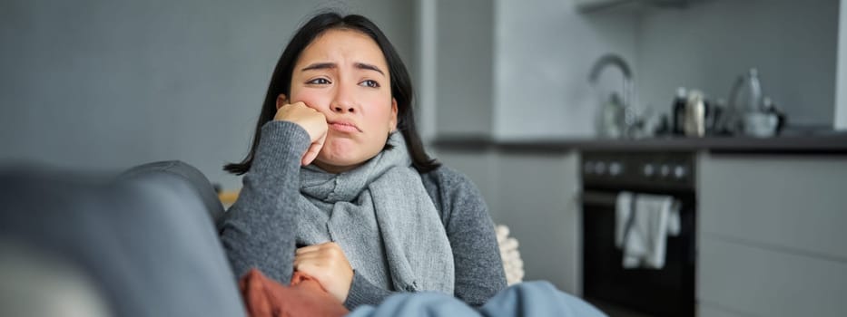 Image of sad young woman feeling sick, being unwell, sitting with scarf and frowning in her living room, catching cold and staying at home on isolation.