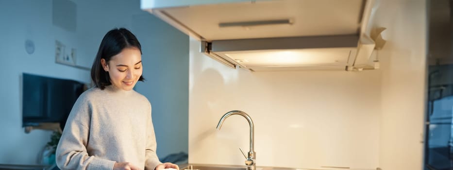 Vertical shot of young asian woman cooking dinner, making herself sandwitch, smiling while standing on the kitchen.