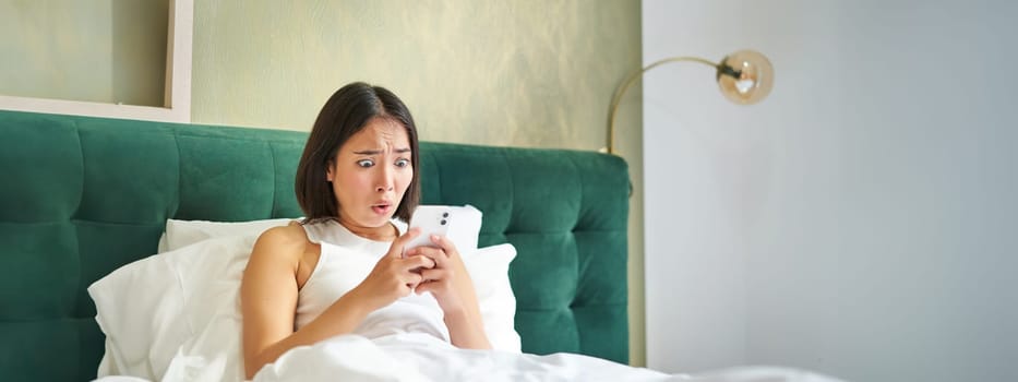 Portrait of shocked korean woman looking at her mobile phone, lying in bed, reading bad shocking news on smartphone app.
