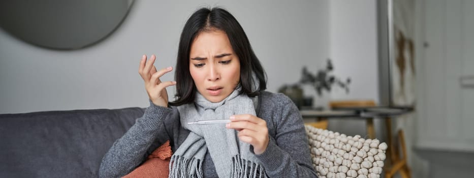 Shocked woman looks concerned at her thermometer, catching cold, has fever, sitting on sick leave at home.