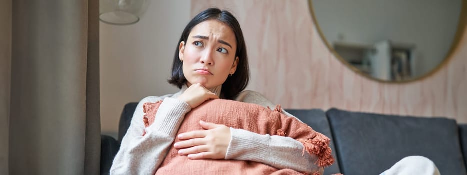 Women and wellbeing concept. Portrait of sad and gloomy asian woman thinking of something unhappy, hugging pillow on sofa and expressing distress.