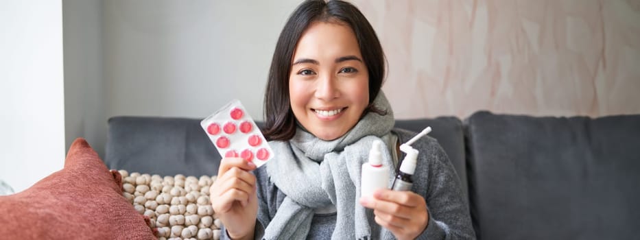 Portrait of happy smiling asian girl showing medication, sore throat spray and drugs from flu or cold, staying at home, wearing warm clothes.
