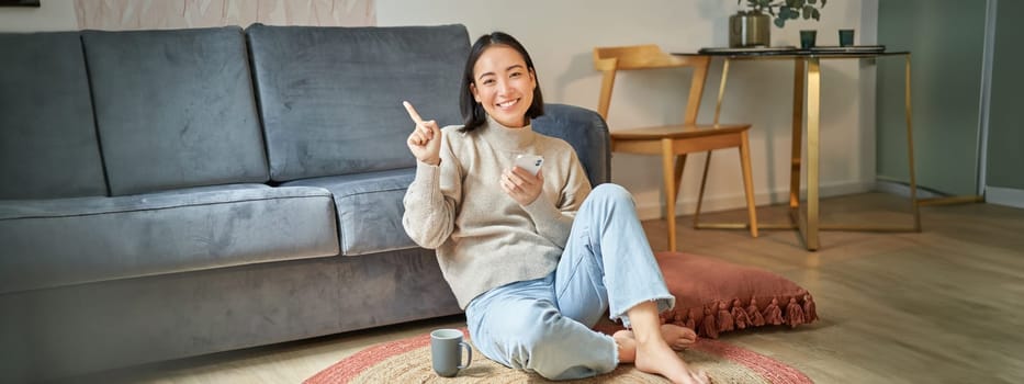 Smiling asian girl sits on floor in stylish living room, pointing finger at advertisement, showing promo banner, holding mobile phone in hand.