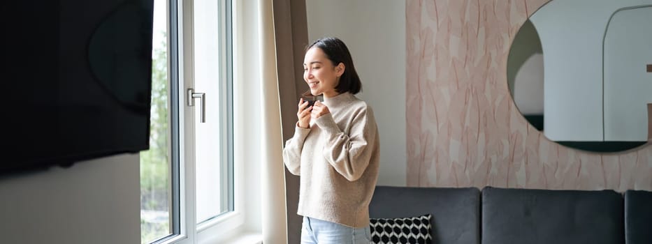 Morning. Happy asian woman drinking espresso at home, looking outside window and smiling, standing in cozy living room feeling warm.