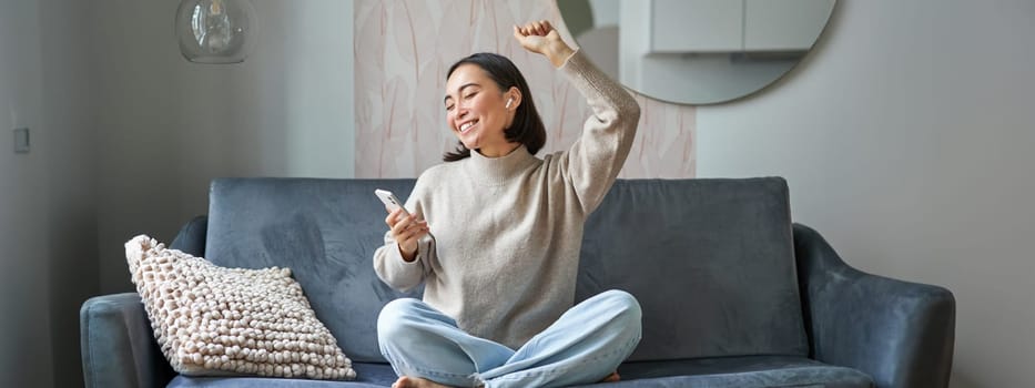 Portrait of carefree asian woman singing and listening music from smartphone app, using wireless headphones, smiling pleased, sitting on sofa at home.