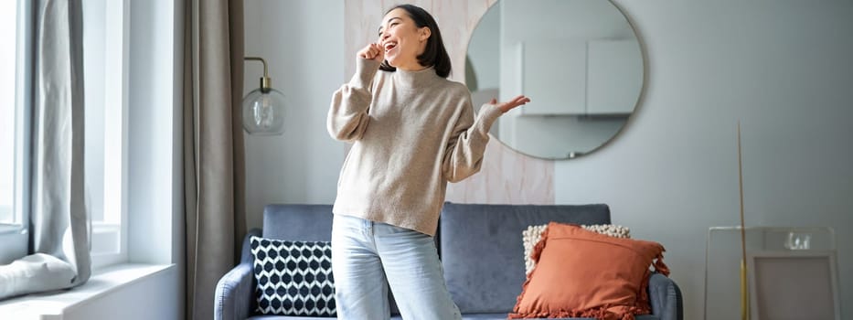 People and emotions. Portrait of asian girl standing in living room, singing in shadow microphone, dancing and having fun, feeling joy.