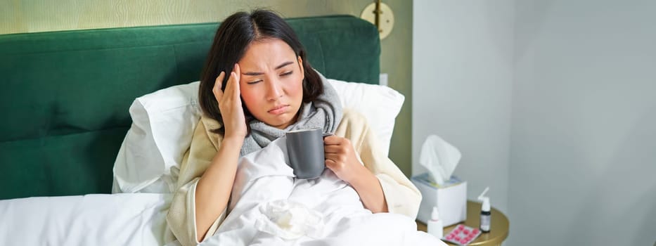 Portrait of asian woman with headache, catching col, staying on sick leave at home, lying in bed, drinking hot tea, having flu.