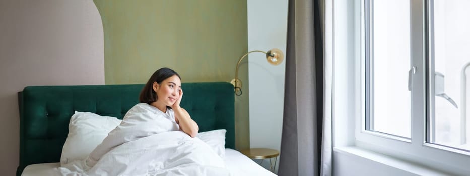 Happy korean woman wakes up in her hotel room, lying in bed under cozy warm blankets, white sheets, enjoying weekend morning.