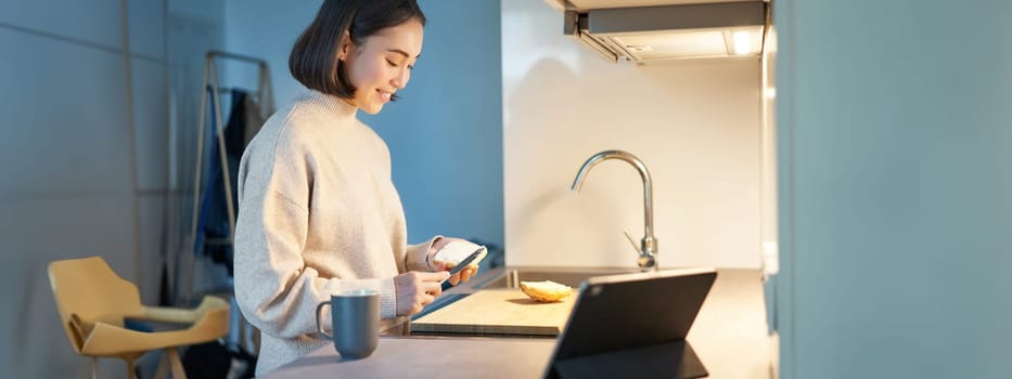 Portrait of young korean woman, making toast and watching video on digital tablet, looking at screen.