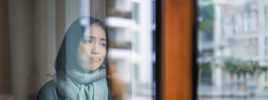 Portrait of sad ill korean woman looking out of the window, staying at home due to coronavirus, being sick and cant leave house.