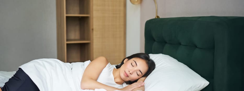 Beautiful asian girl sleeping in her bed, lying in bedroom with closed eyes under white sheets, warm winter duvet.