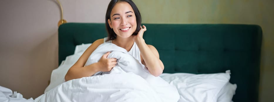 Close up of happy beautiful asian woman, waking up in bed and enjoys morning, looking outside window with sleepy smile on her face, hugging duvet.