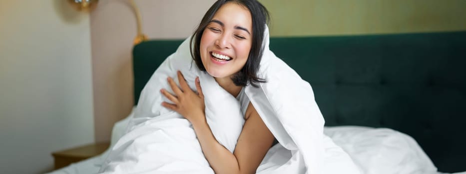 Beautiful asian woman sitting on bed, covered with white duvet, smiling, enjoying happy weekend morning, laughing at camera.