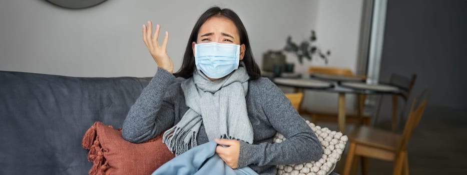 Covid-19 and health concept. Young asian woman in medical face mask, feels sick and unwell, catching flu, protecting others from influenza, sitting in living room covered with blanket.