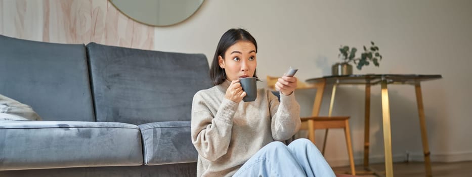 Portrait of young korean woman watching television, holding remote and looking amazed at tv screen, spending time at home.