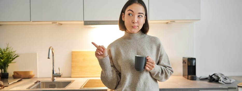 Portrait of smiling asian girl standing in kitchen, drinking coffee from cup and pointing at banner, showing letting agencies advertisement.