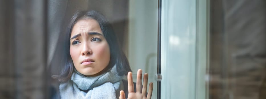 Vertical shot of upset asian woman looking outside, touching window, staring at street with gloomy jealous face, feeling ill, staying at home with covid or flu.