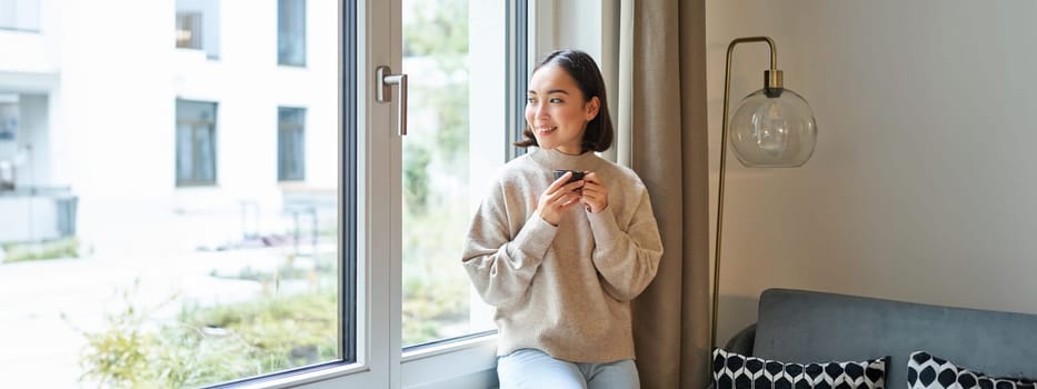 Beautiful young asian woman sitting near window and drinking her coffee, holding espresso cup and looking outside with relaxed, smiling face expression.