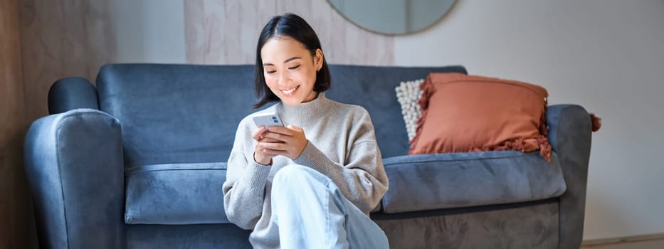 People and technology. Smiling asian woman sitting at home, using her mobile phone, typing message, browing internet or shopping online from smartphone app.