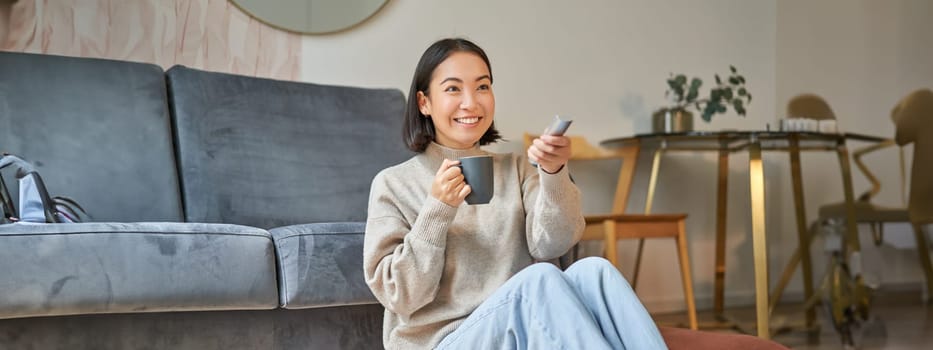 Portrait of young korean woman watching television, holding remote and looking amazed at tv screen, spending time at home.