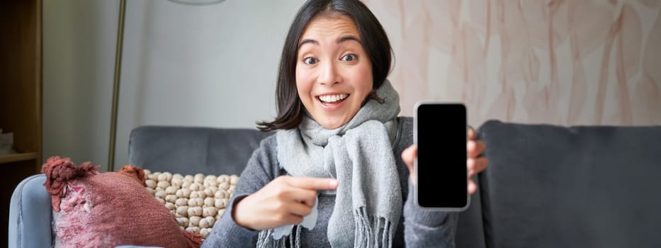 Excited young woman pointing finger at smartphone, showing online doctor, medical application or GP contact on mobile phone, staying at home sick, catching cold.