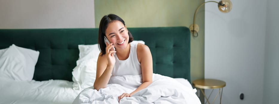 Beautiful young korean woman calling, having a phone call in bed, talking with friend while staying in bedroom.